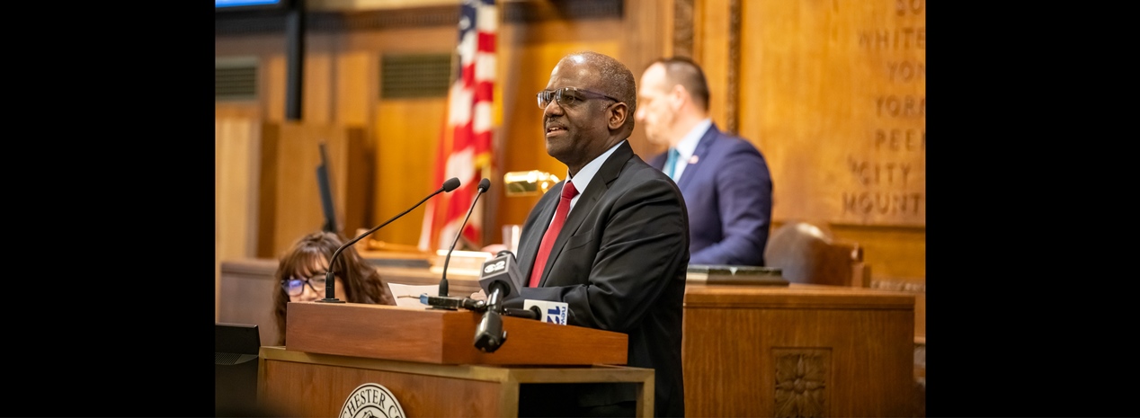 A man wearing glasses, a black suit, a white shirt, and a red tie speaks at a podium in a formal setting. Behind him are an American flag, microphones, and other individuals seated in the background. The setting appears to be a government chamber or courtroom.