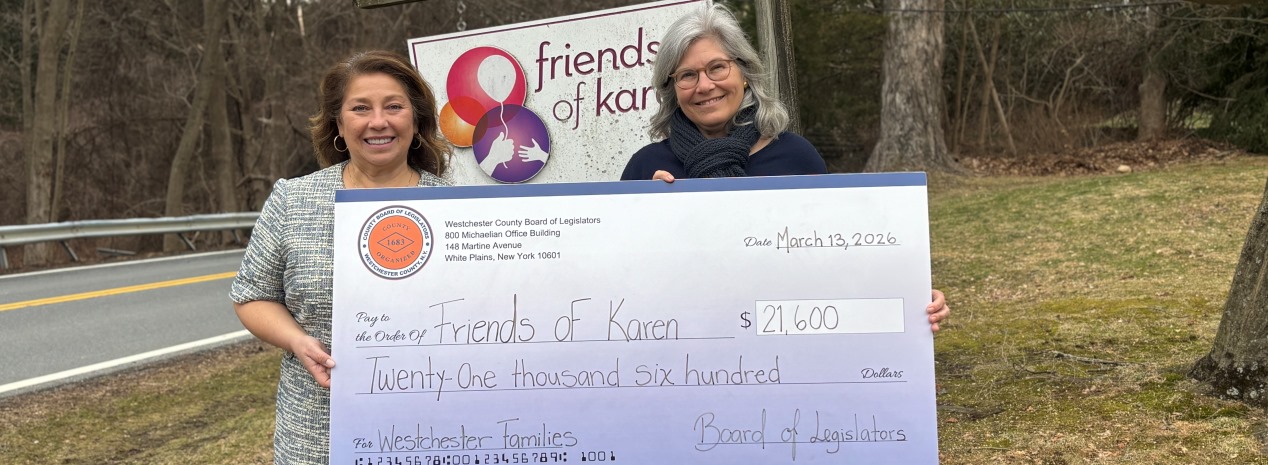 Legislator Pierce stands outdoors holding an oversized ceremonial check with another individual in front of a Friends of Karen sign. 