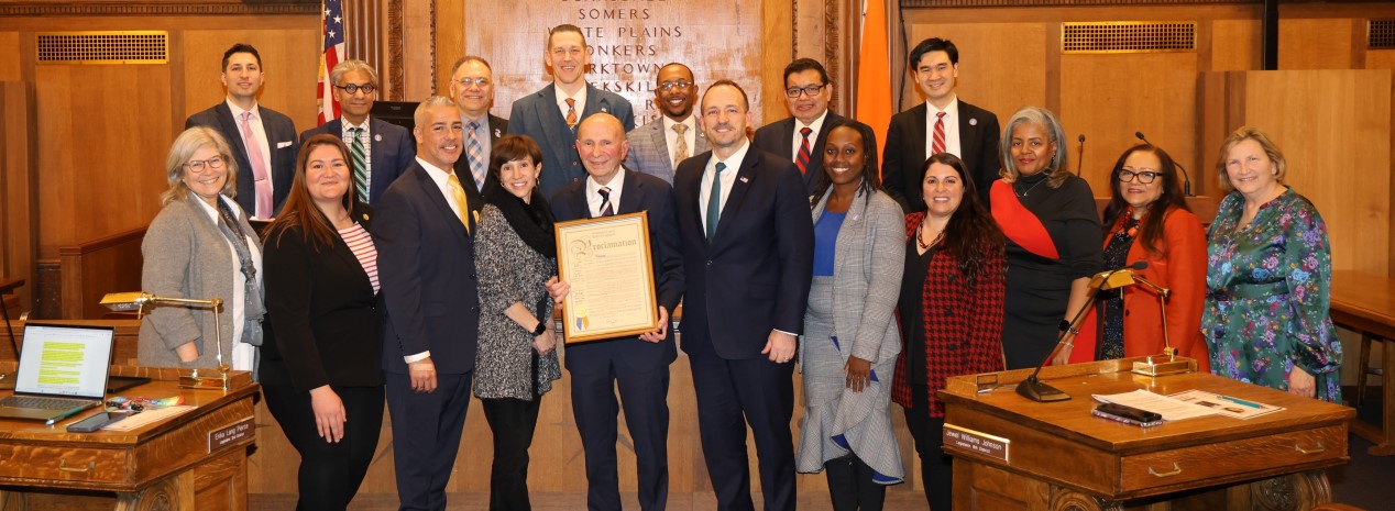Group photo of Westchester County legislators standing in the legislative chamber, gathered around Richard Wishnie holding a framed proclamation.